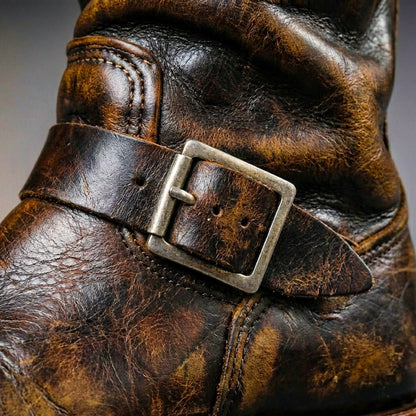 Close-up of the ankle strap and square antique-silver buckle on a scuffed, creased brown leather boot. 5