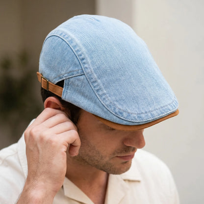 Man in a cream shirt wearing the light blue denim flat cap, head tilted down, tan suede-look peak and buckle strap visible. 2