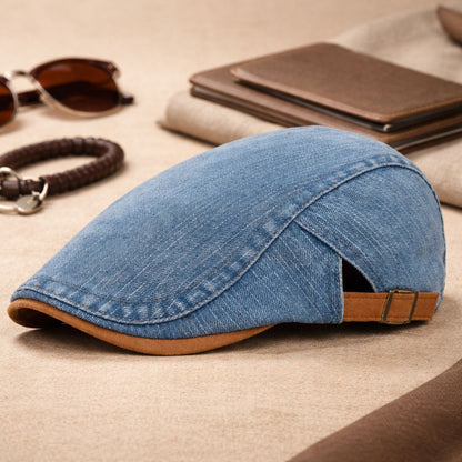 Mid-blue denim flat cap in a side view on a beige surface, surrounded by sunglasses, a bracelet and wallets. 6