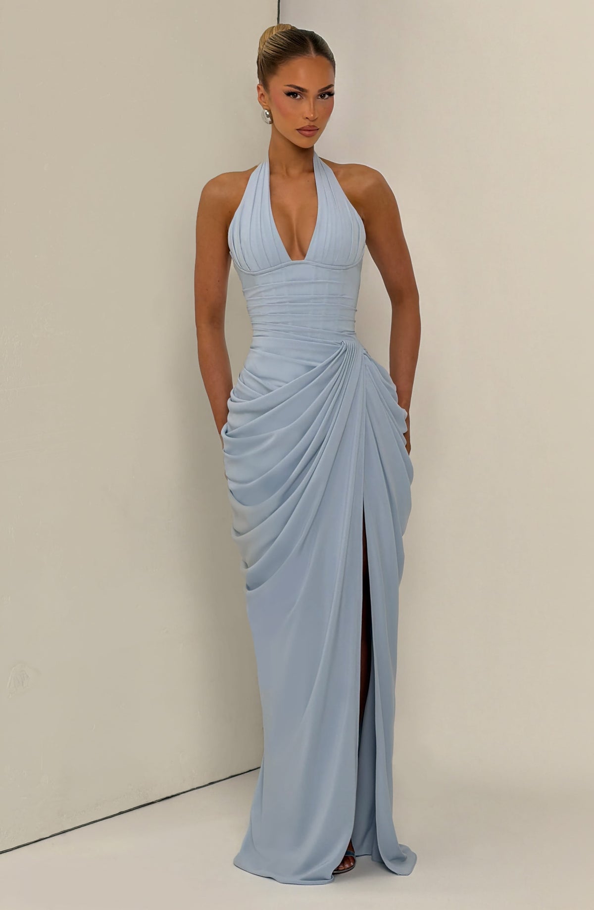 Pale blue dress on a model leaning against a cream wall, with her hands by her sides and a soft slit in the skirt. 15