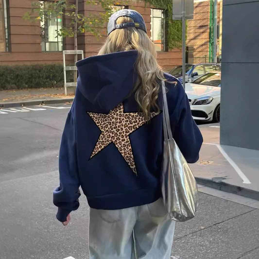 Back view of a woman on a street wearing the navy hoodie with its large leopard-print star, with a cap and silver shoulder bag. 1