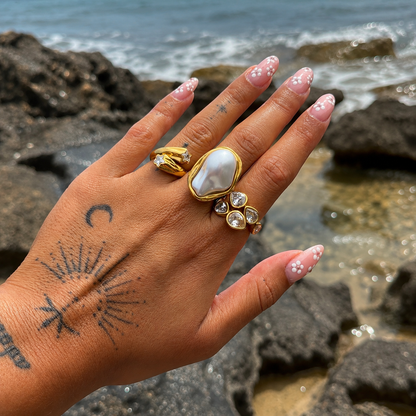 Hand held up before rocks and sea, wearing the gold star ring next to a large pearl-style ring and a teardrop crystal ring. 5