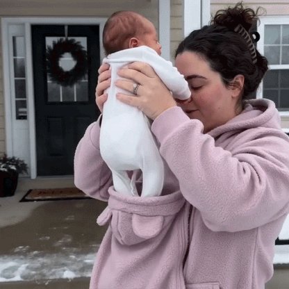 Woman in the pink fleece hoodie lifting a baby in white out of the front pouch outside a front door. 11