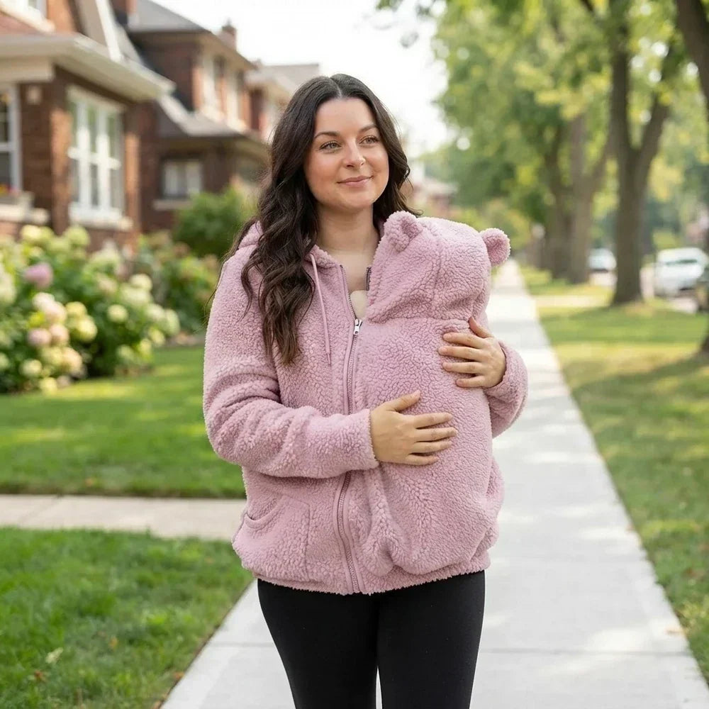 Woman in the pink fleece hoodie walking down a leafy street with the bear-eared pouch zipped over her baby. 15