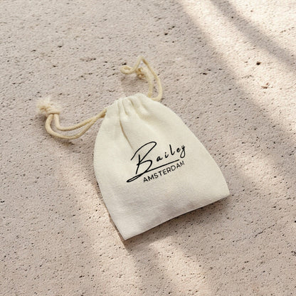 Cream cotton drawstring pouch with a black printed logo, lying on a sandy textured surface in dappled sunlight. 7