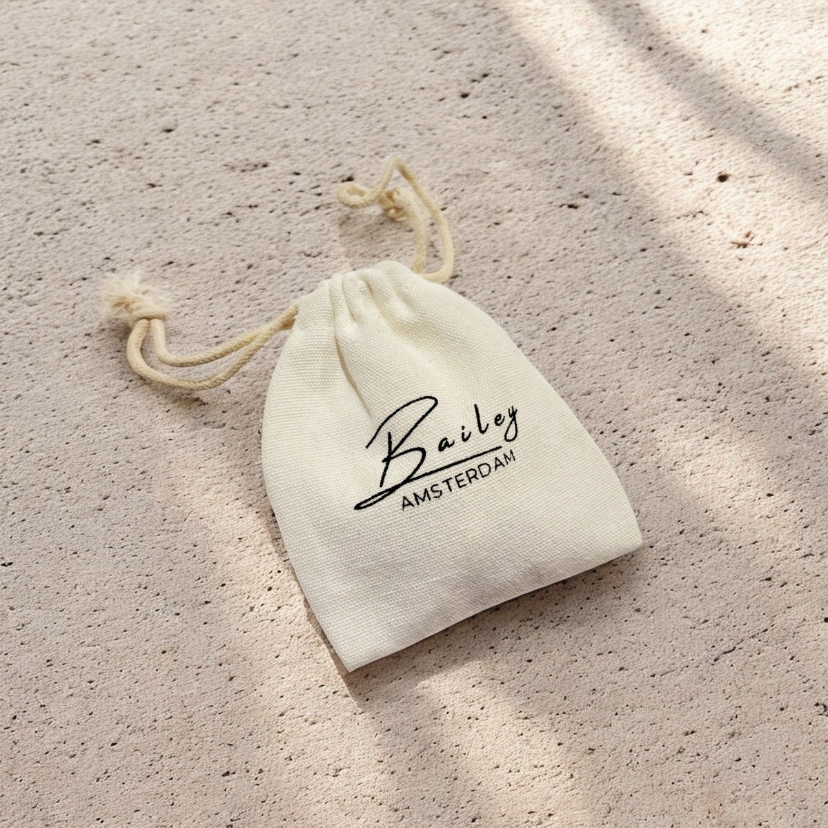 Small cream cotton drawstring pouch with a black printed logo, lying on a sandy textured surface in dappled sunlight. 6