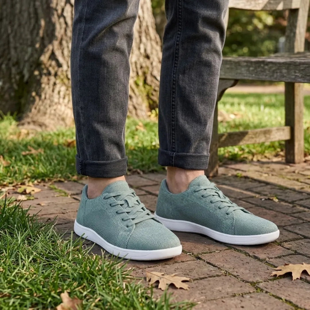 Person wearing sage green knit trainers with white soles and rolled grey jeans, standing on brick paving near a bench. 29