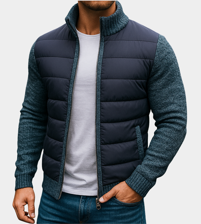 Man wearing the quilted jacket in navy with blue-grey knitted sleeves and collar, one hand in his jeans pocket. 4
