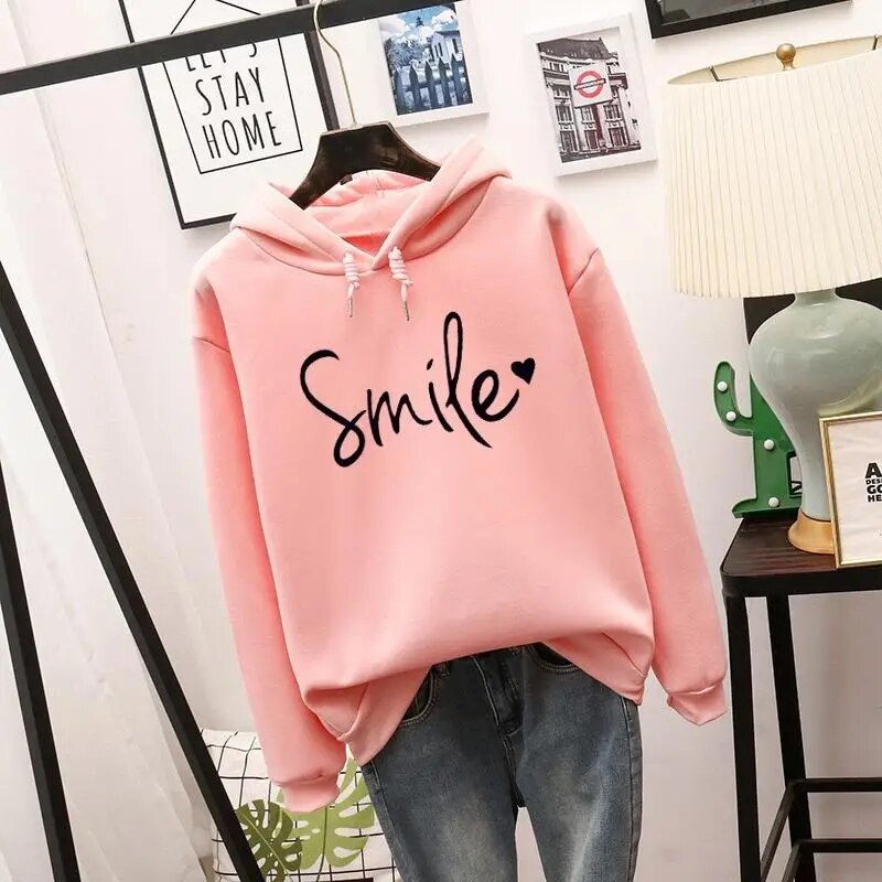 Pink version of the hooded sweatshirt with the same black script slogan and heart, hanging on a hanger over blue jeans. 2
