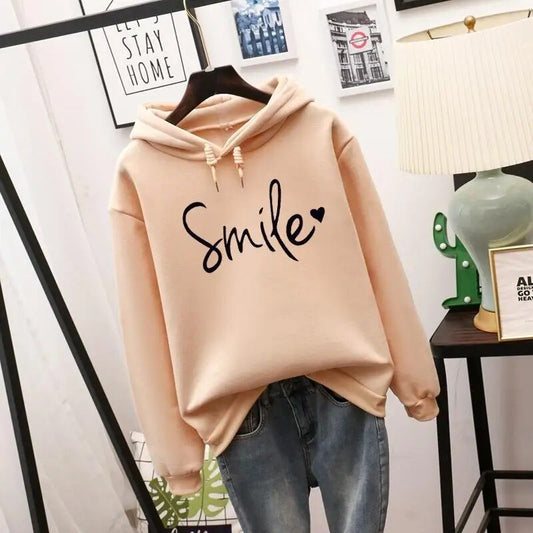 Beige hooded sweatshirt with drawstrings and a black heart-dotted script slogan across the chest, hung on a hanger above blue jeans. 1