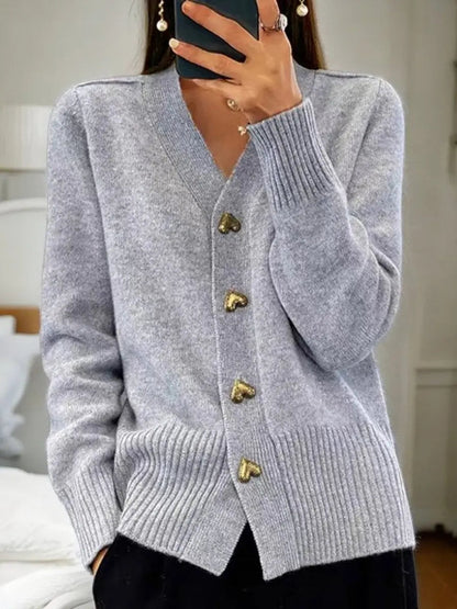 Light grey version of the cardigan worn by a model, showing gold heart buttons, V-neckline and ribbed hem with black trousers. 2
