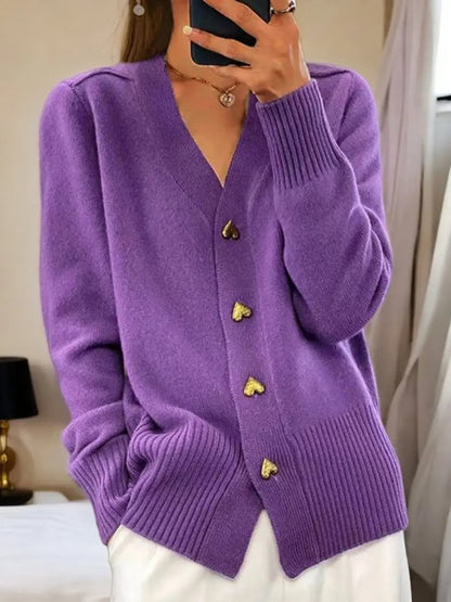 Purple version of the cardigan on a model, with four gold heart buttons, ribbed cuffs and hem, worn with white trousers. 3