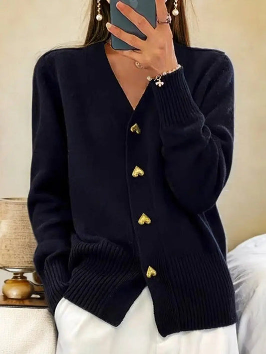 Woman taking a mirror selfie in the navy knit cardigan with gold heart buttons, ribbed cuffs and hem, worn over white trousers. 1