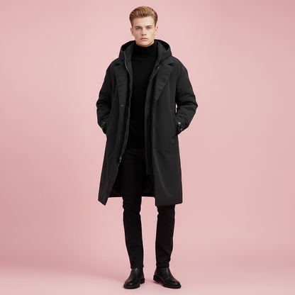 Model standing front-on in the black hooded coat, hands in pockets, worn open over black roll-neck, trousers and boots against pink. 3