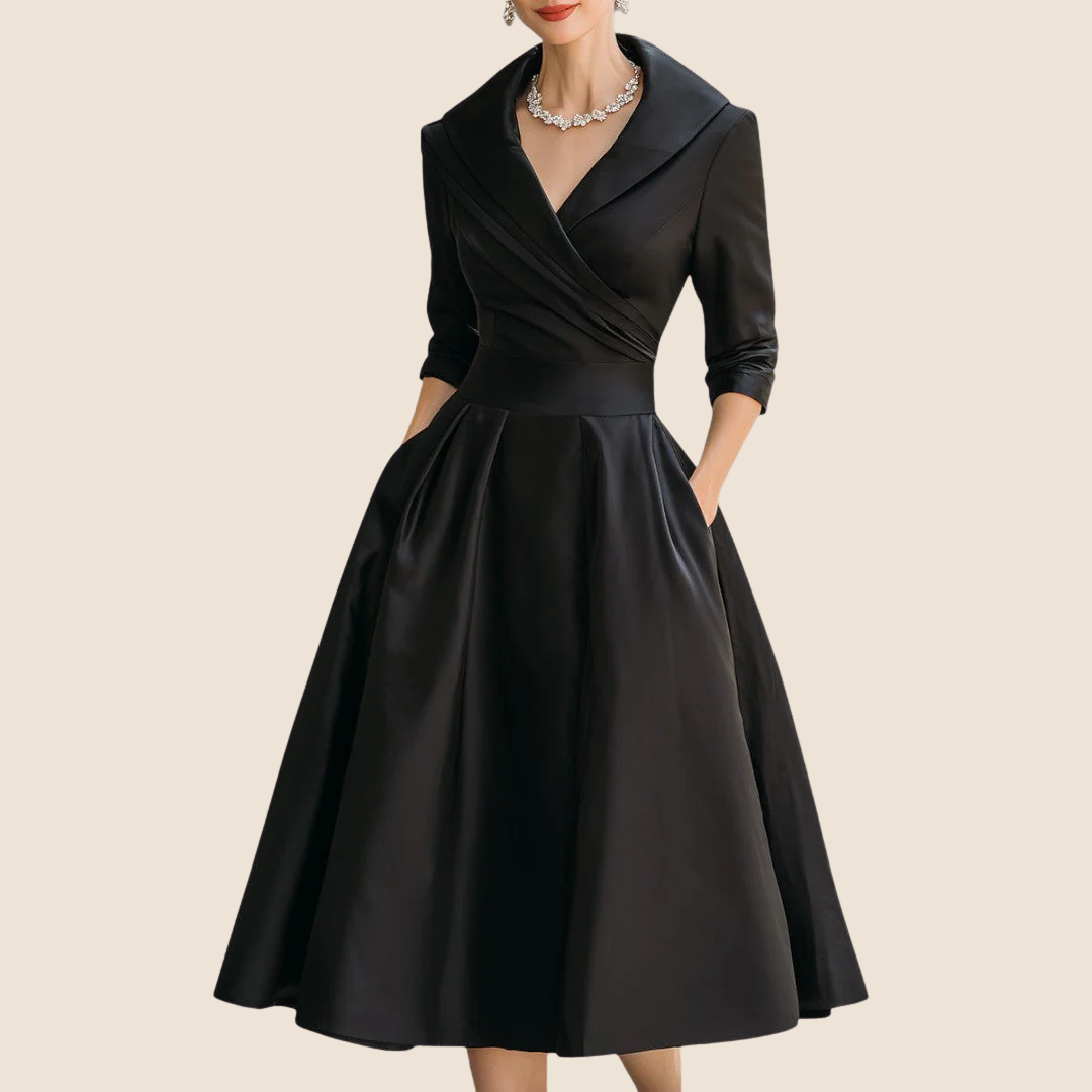 Model wearing the black satin A-line midi dress with shawl collar, three-quarter sleeves and pockets, styled with a crystal necklace. 1