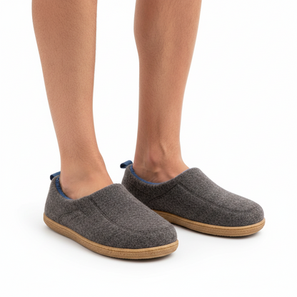 Bare legs wearing the charcoal grey slippers, with blue lining and heel loops visible and tan soles on white background. 10