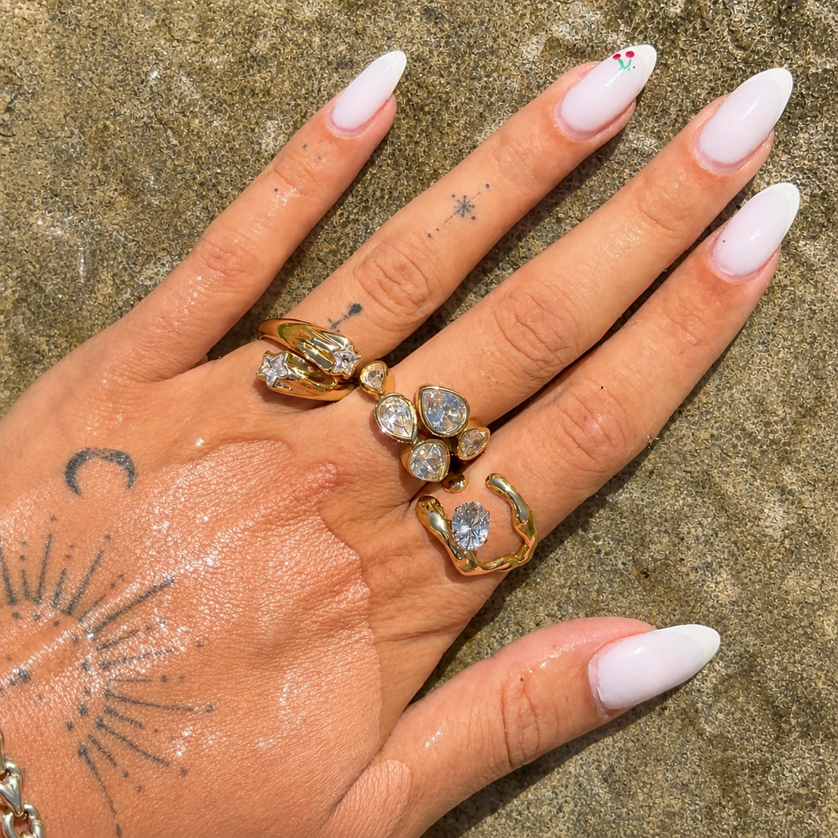 Hand with white manicured nails wearing the gold star ring on one finger, stacked with other crystal rings, resting on a sandy surface. 3