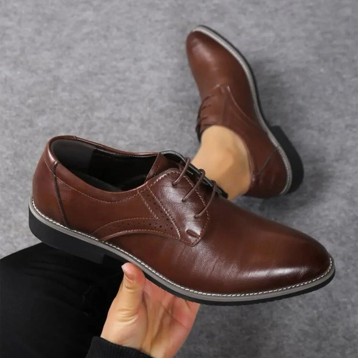 Dark brown polished lace-up shoe held in a hand from above, with a second shoe worn on a foot behind. 2
