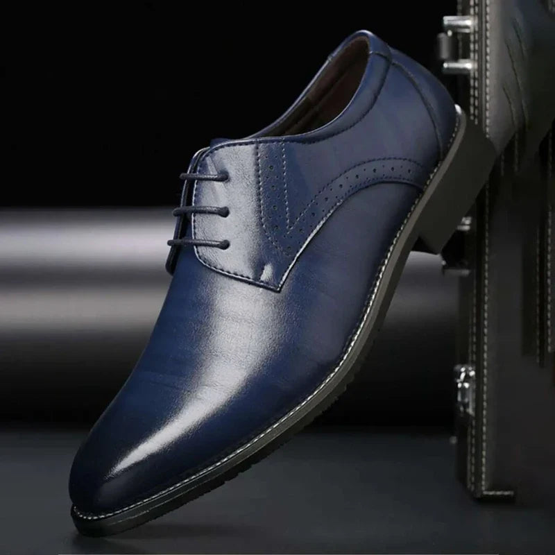 Navy blue polished lace-up shoe with brogue perforations and a pointed toe, displayed side-on beside a dark briefcase. 5