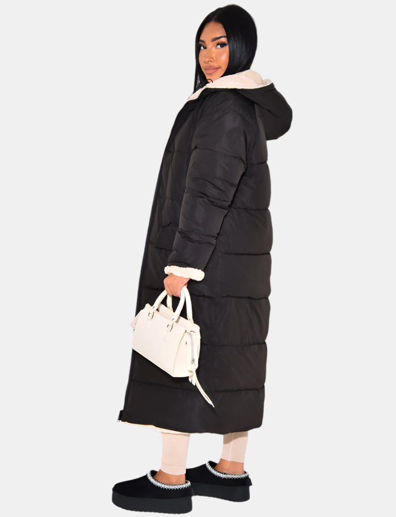 Side view of the black quilted coat worn zipped to the ankle, with a fleece-trimmed cuff and hood, holding a white handbag. 6