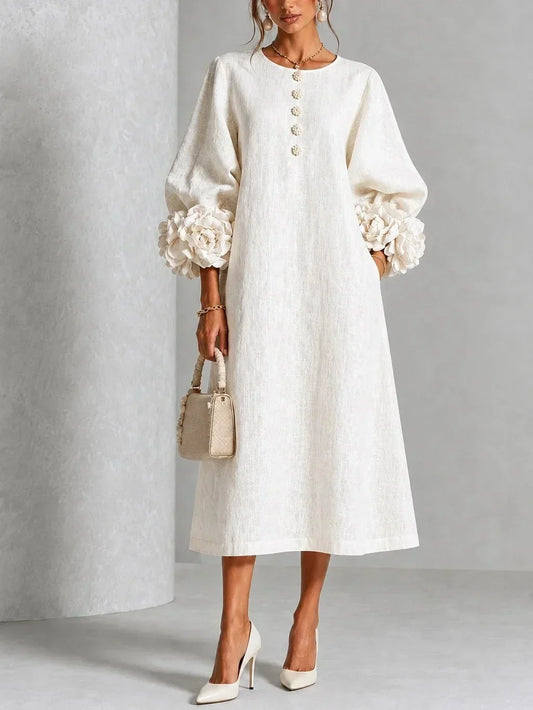 Model wears the cream long-sleeved midi dress with oversized fabric flowers at the cuffs and flower buttons down the front, holding a woven handbag. 1