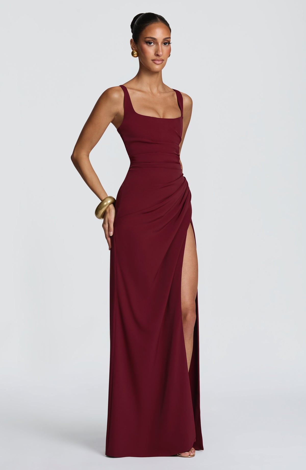 Angled view of the burgundy dress with one hand on the hip, showing ruched hip and high slit. 19