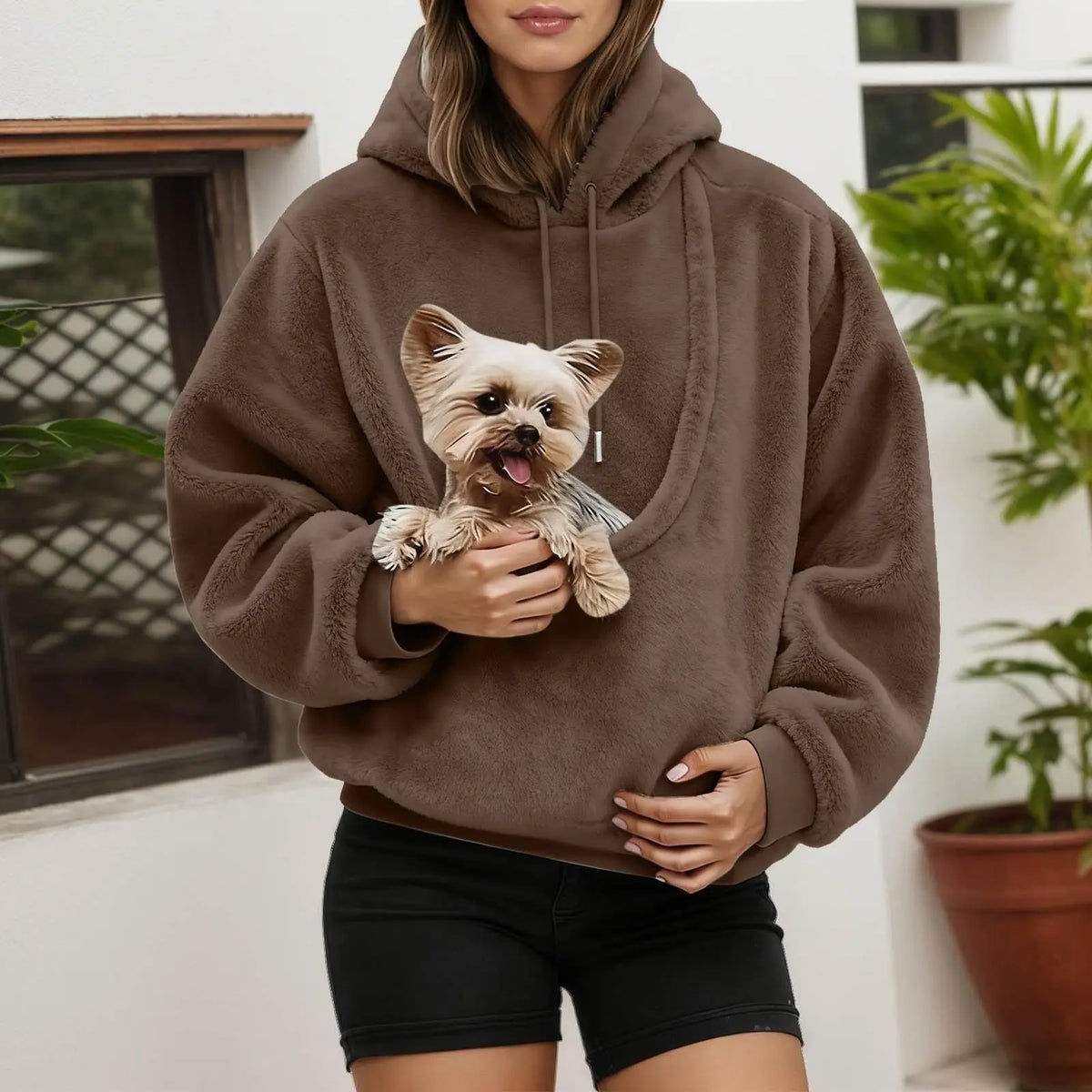 Woman wearing a brown fluffy hoodie beside plants, cradling a small terrier in the front pouch. 3