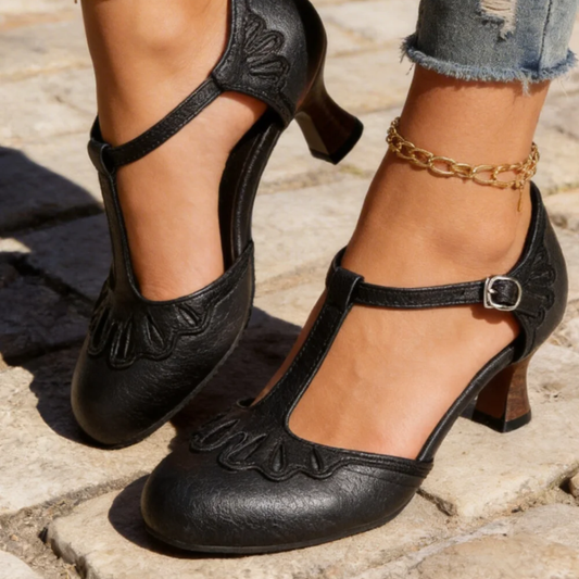 All-black version of the T-bar pumps worn outdoors, showing the textured finish, scalloped overlay and wooden-look block heel. 2