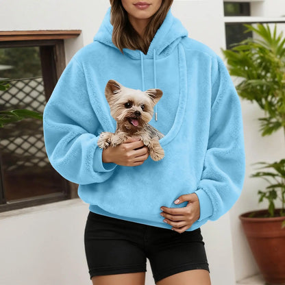 Woman wearing a light blue fluffy hoodie, holding a small terrier in the front pouch beside a plant. 8
