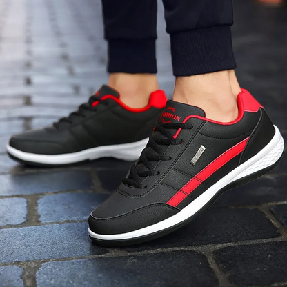 Black sneakers with red side stripe and white sole worn by a man in dark joggers on cobbled paving. 2