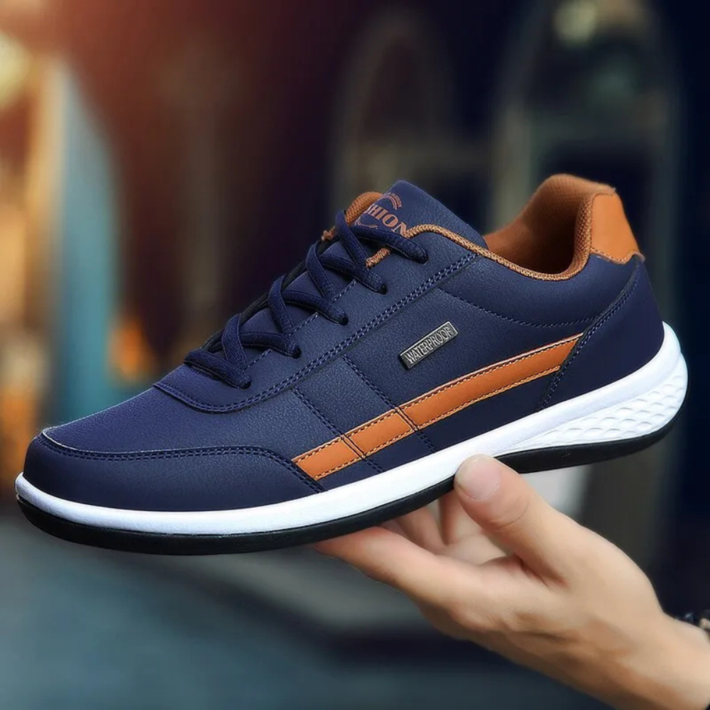 Navy blue sneaker with tan side stripe and white sole held in an outstretched hand against a warm blurred backdrop. 6