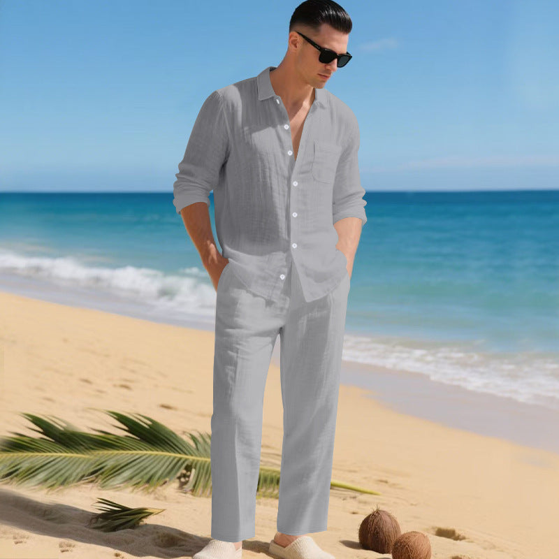 Man in sunglasses on a sandy beach wearing the light grey long-sleeved shirt and matching straight trousers, hands in pockets. 2