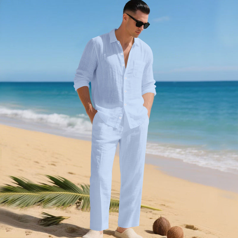 Man in sunglasses on a sandy beach wearing the pale blue long-sleeved shirt and matching straight trousers, hands in pockets. 3