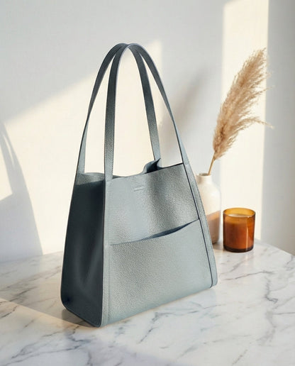 Light blue-grey leather tote with long straps and a front pocket on a marble surface beside pampas grass and an amber candle. 10