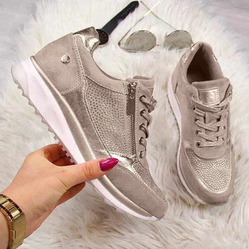 Hand holding the champagne trainers over a fluffy rug, showing the side zip, studded panels and chunky white sole. 7