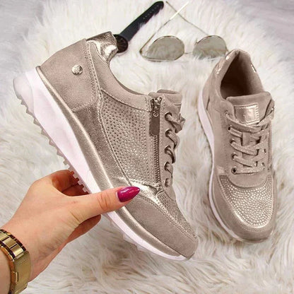Hand holding the champagne trainers over a fluffy rug, showing the side zip, studded panels and chunky white sole. 7