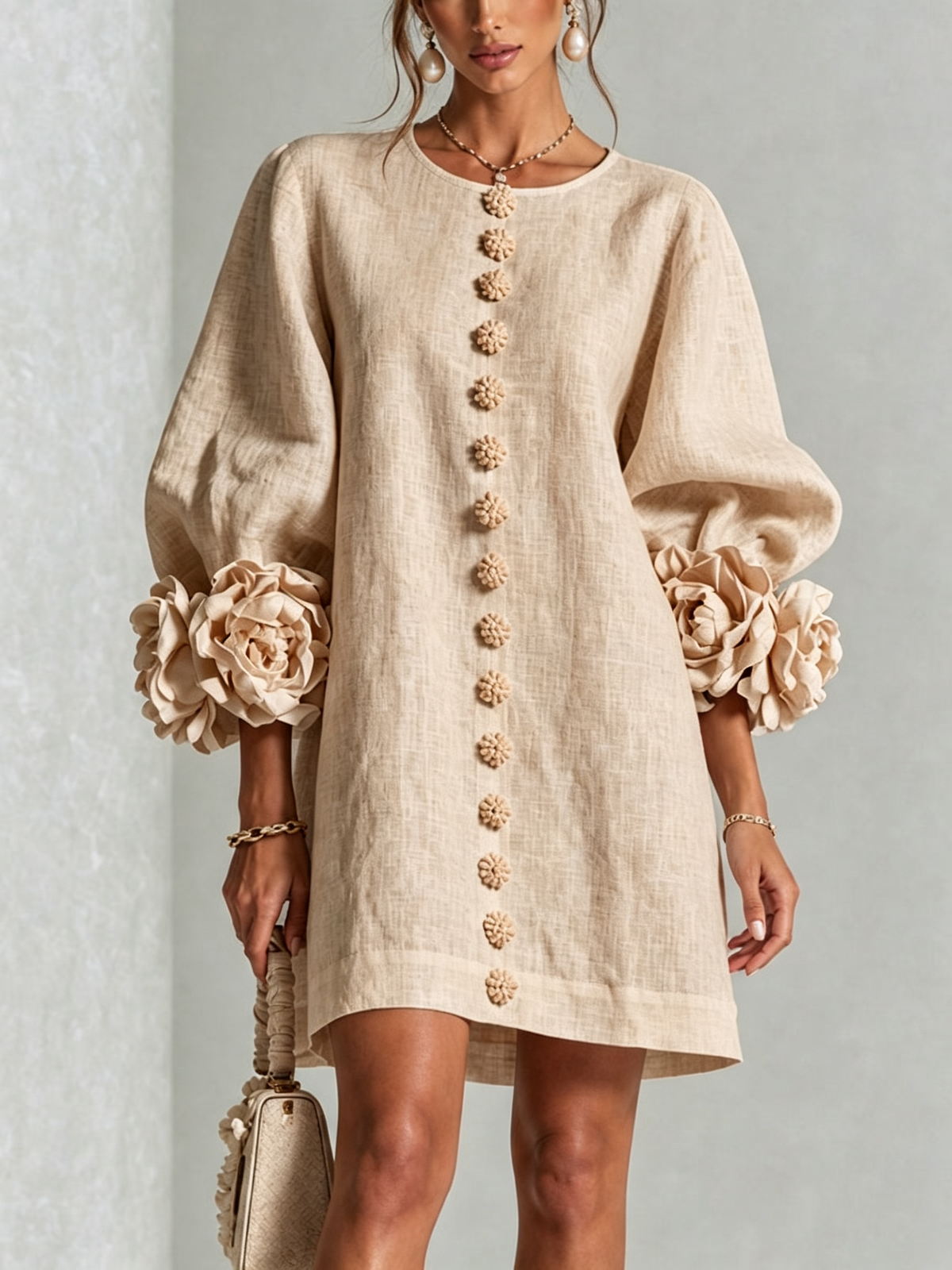 Beige linen-look dress worn by a model, front view, with fabric flower buttons down the front and rosette-trimmed puffed sleeves. 27