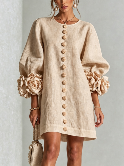 Beige linen-look dress worn by a model, front view, with fabric flower buttons down the front and rosette-trimmed puffed sleeves. 27