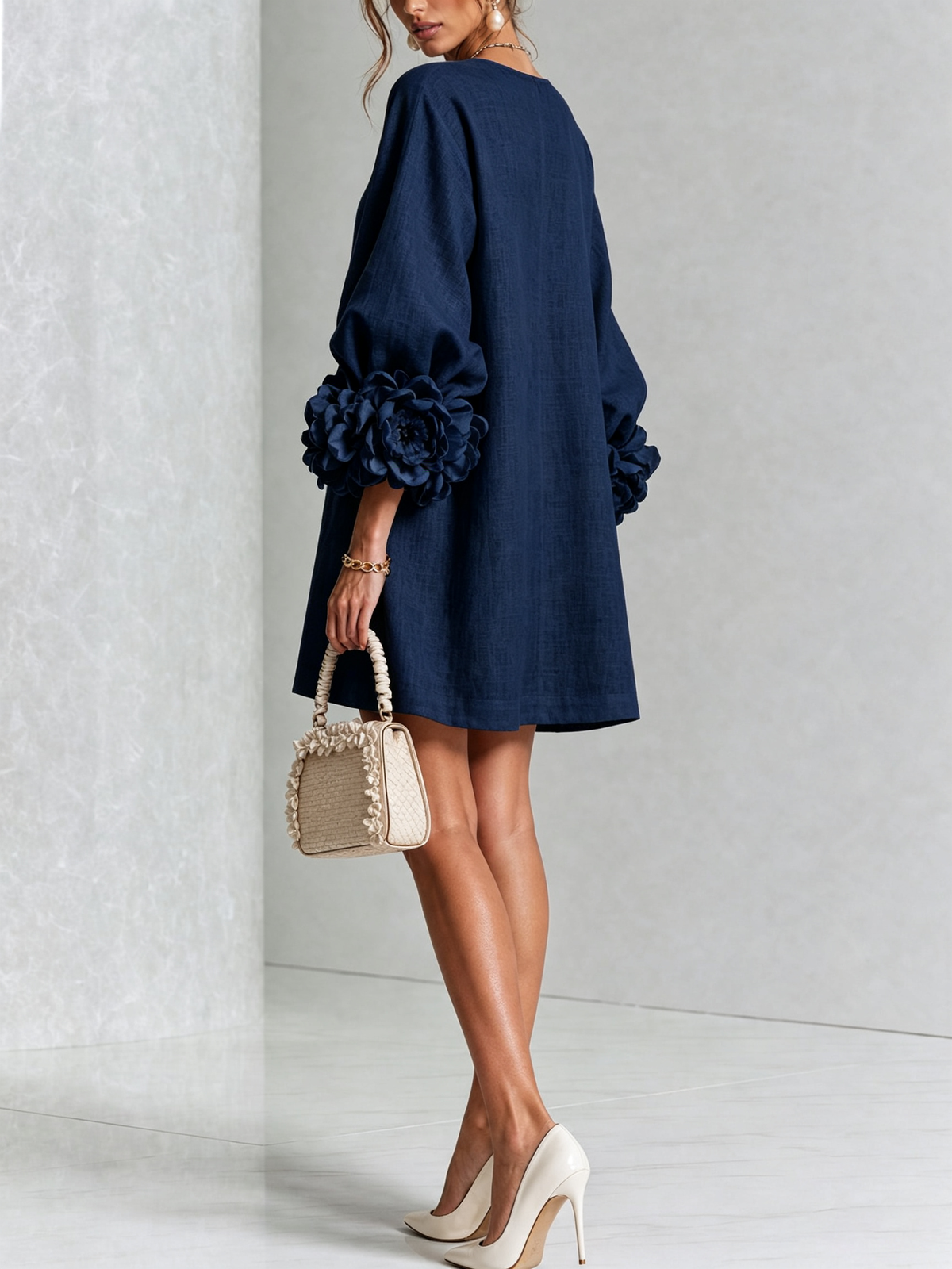 Back view of the navy dress in a studio, showing the plain back, rosette cuffs on balloon sleeves, and a woven handbag. 20