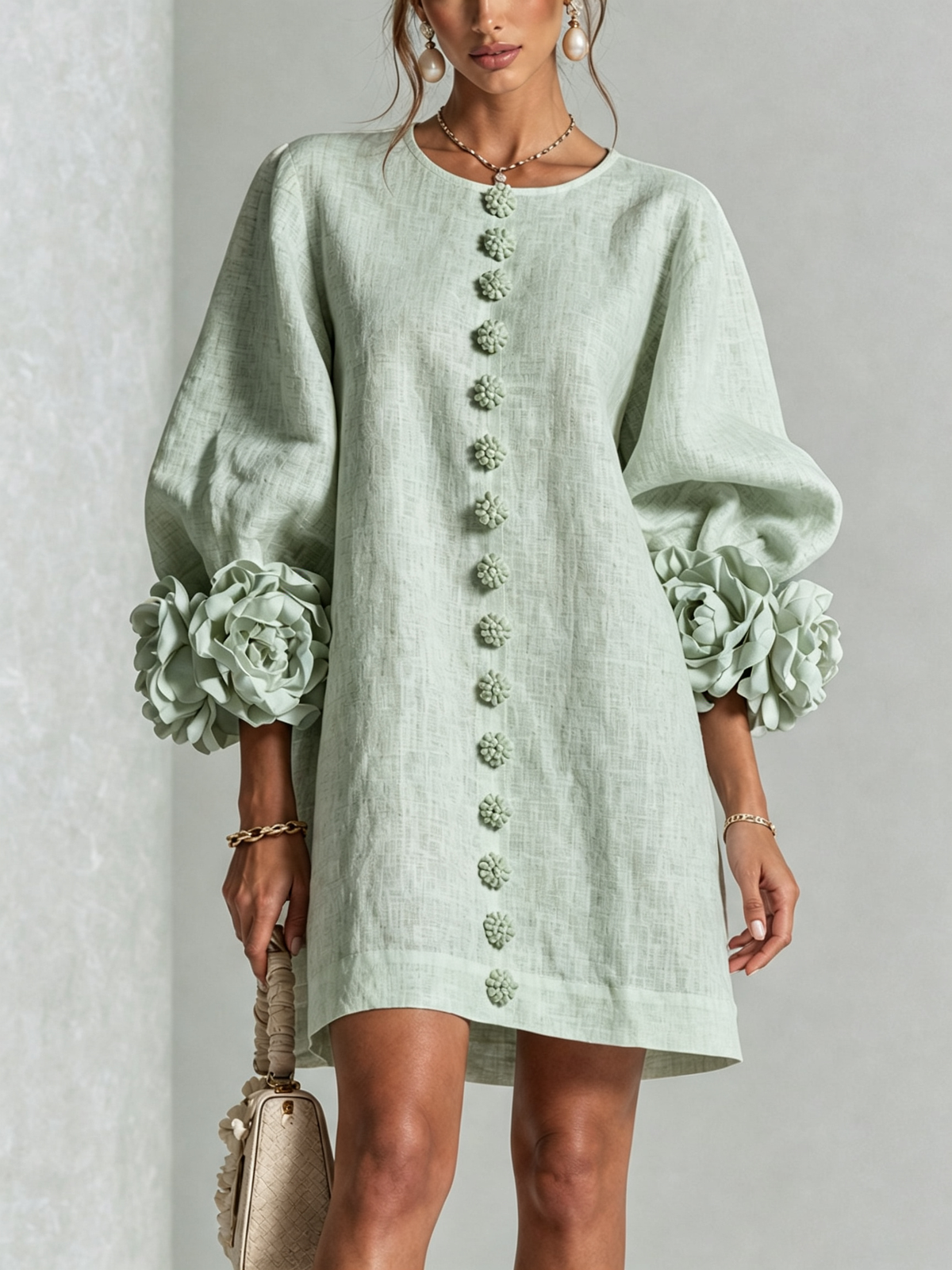 Pale sage green dress worn by a model, front view, with fabric flower buttons down the front and large rosettes at the sleeve cuffs. 35
