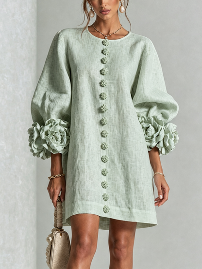 Pale sage green dress worn by a model, front view, with fabric flower buttons down the front and large rosettes at the sleeve cuffs. 35