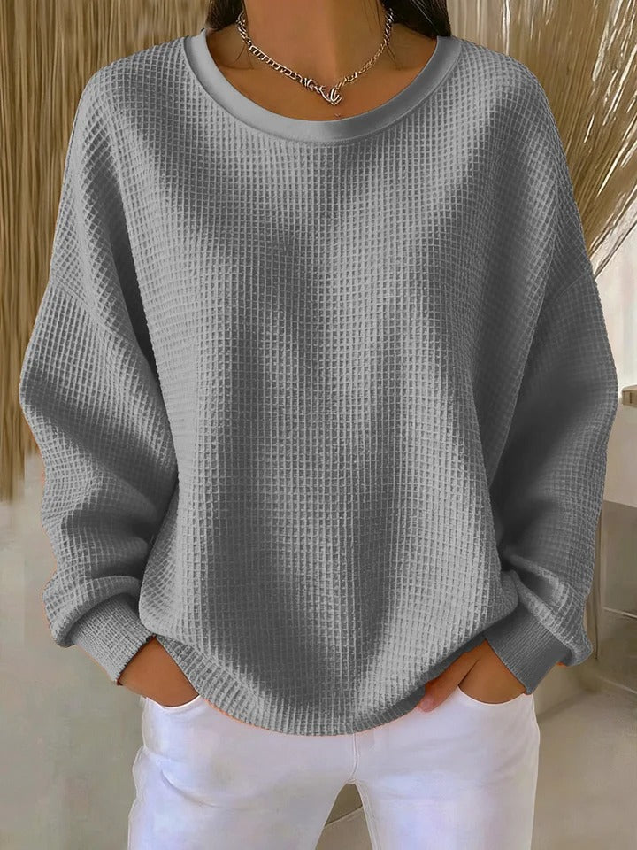 Model wearing the waffle-knit jumper in grey, hands tucked into white trouser pockets, with a silver chain necklace. 3