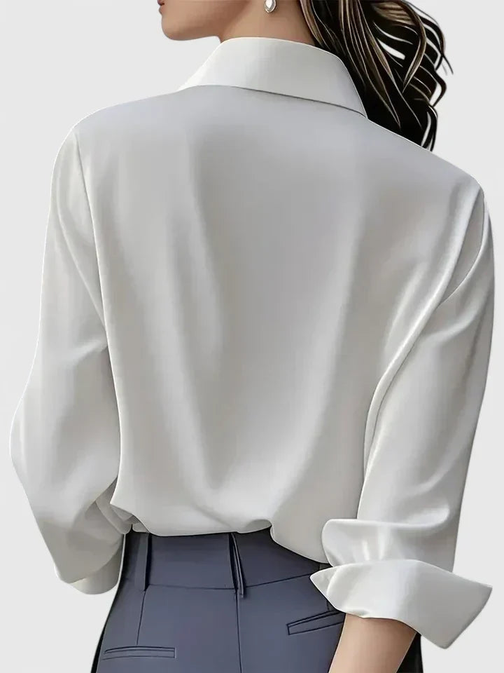 Back view of the white blouse on a model, showing the smooth back, turned-up cuff and collar, worn with grey trousers. 7