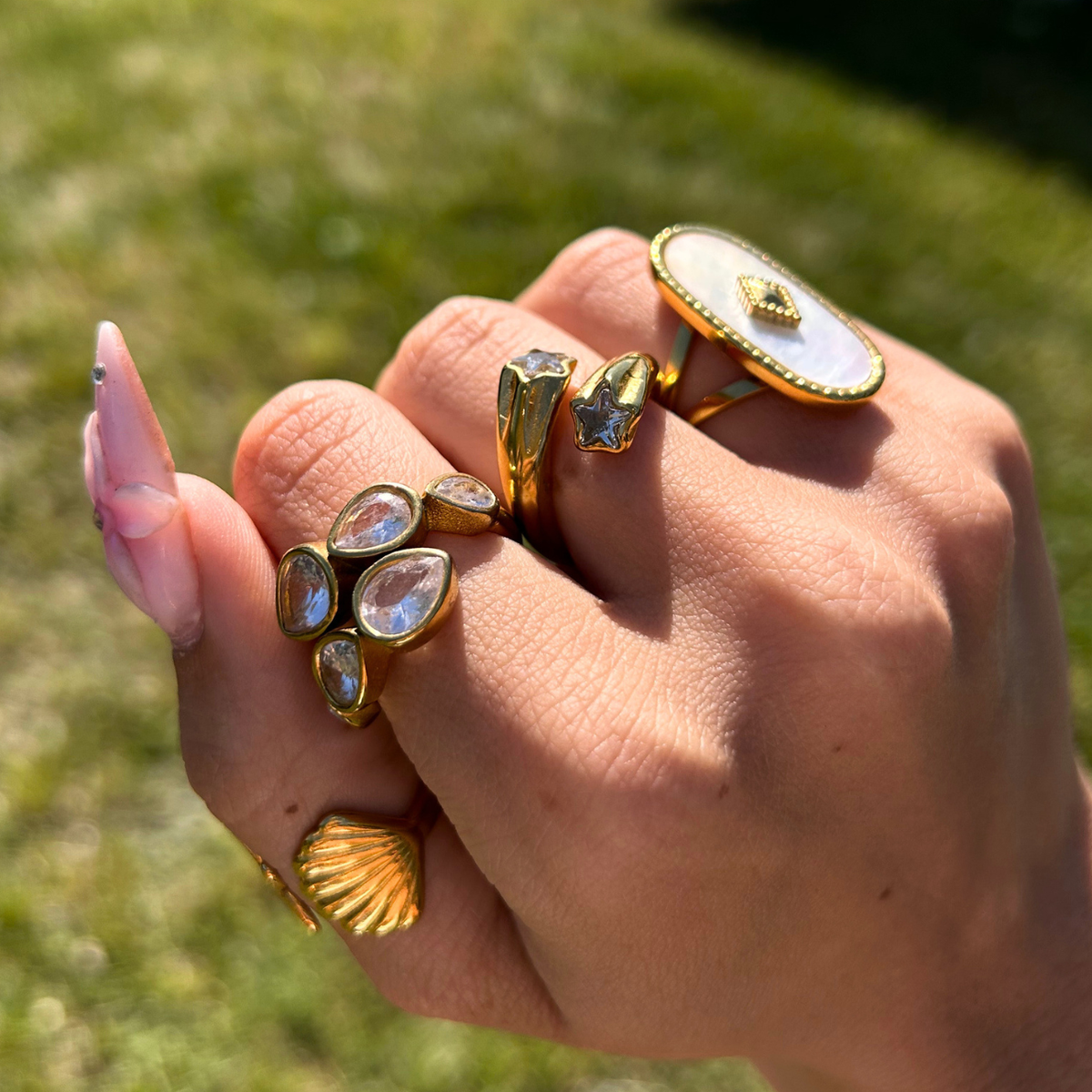 Hand outdoors on a grassy background wearing the star ring alongside other gold rings, including a teardrop crystal ring and an oval pearly one. 4