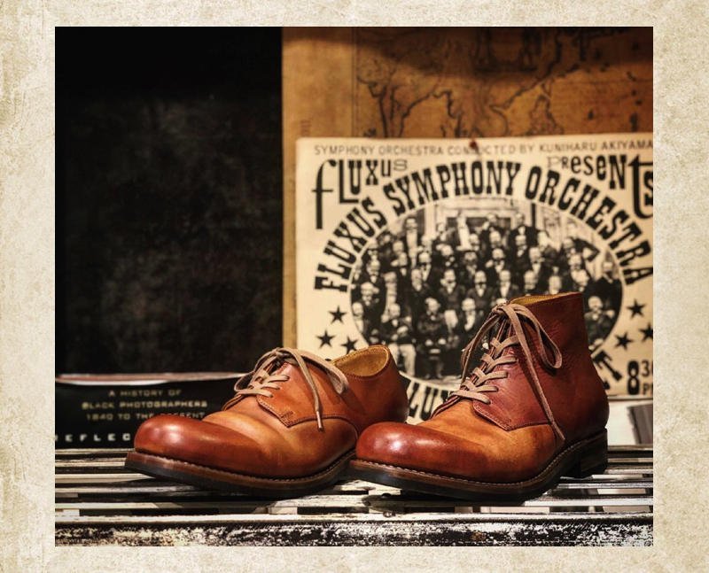 Brown leather lace-up boots with worn, glossy toes standing on a slatted wooden shelf against a vintage poster backdrop. 3