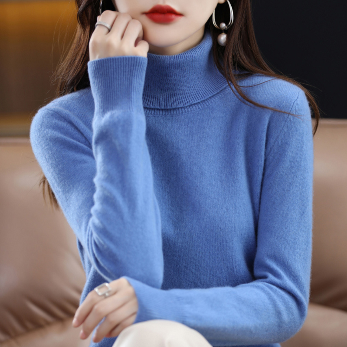 Model wearing the roll neck jumper in bright blue, one hand raised to her chin, seated on a tan sofa. 3