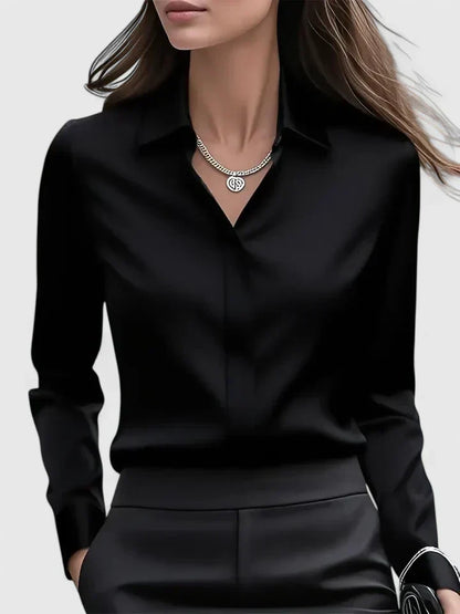 Front view of the black satin blouse on a model, open at the collar with a silver pendant necklace, worn with black trousers. 3