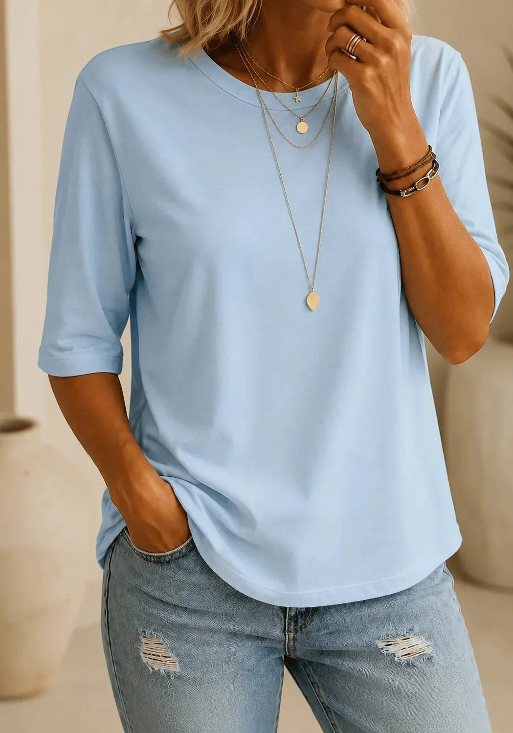 Woman wearing a light blue half-sleeve round neck tee with layered gold necklaces and distressed light-wash jeans, one hand in her pocket. 1