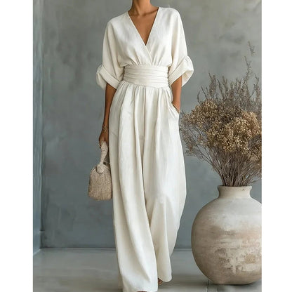 Cream wide-leg jumpsuit with a wrap V-neck and gathered waistband, worn against a grey wall beside a large vase of dried flowers. 1