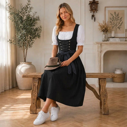 Model sits on a rustic wooden bench in the black dirndl, holding a brown felt hat with a feather in her lap. 3